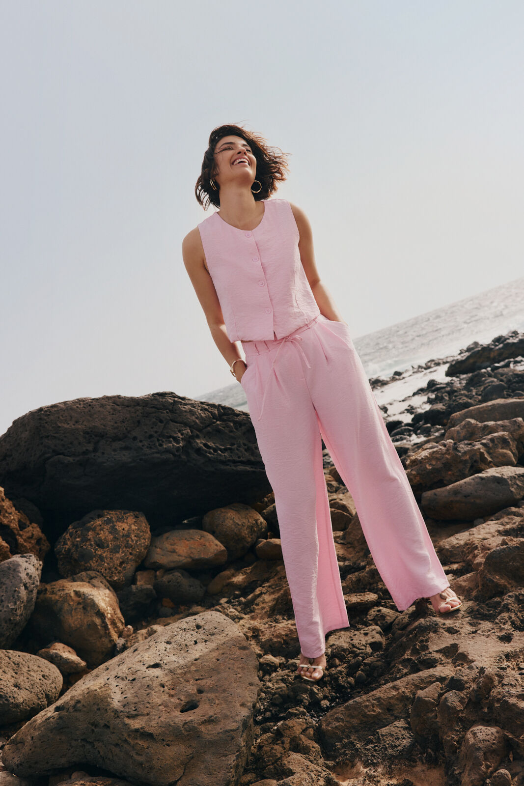 Woman in a pink sleeveless top and matching trousers standing on rocky ground by the sea.